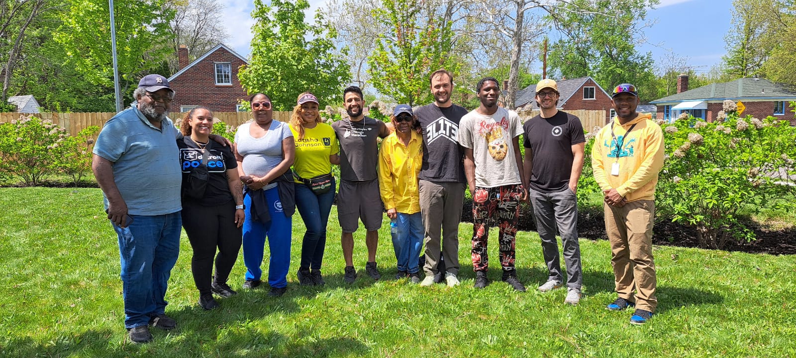 Ten people stand shoulder to shoulder in the sunshine, surrounded by blooming bushes and freshly shorn grass. The people embrace each other and smile at the camera.